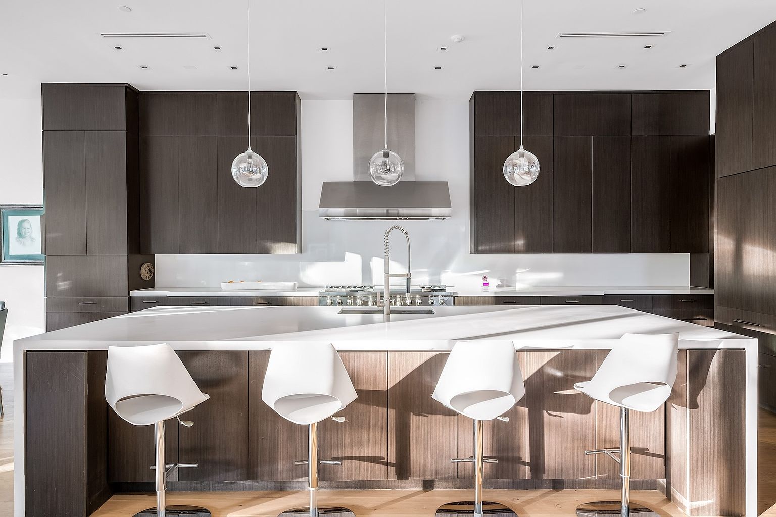 This modern kitchen features dark, flat-panel cabinetry contrasted by a large, light-toned island with seating for four. A stainless steel range hood sits above the cooktop, flanked by glass pendant lights that hang from the ceiling. The space is bright and minimalist, with clean lines and a neutral color palette that creates a sophisticated, contemporary atmosphere.