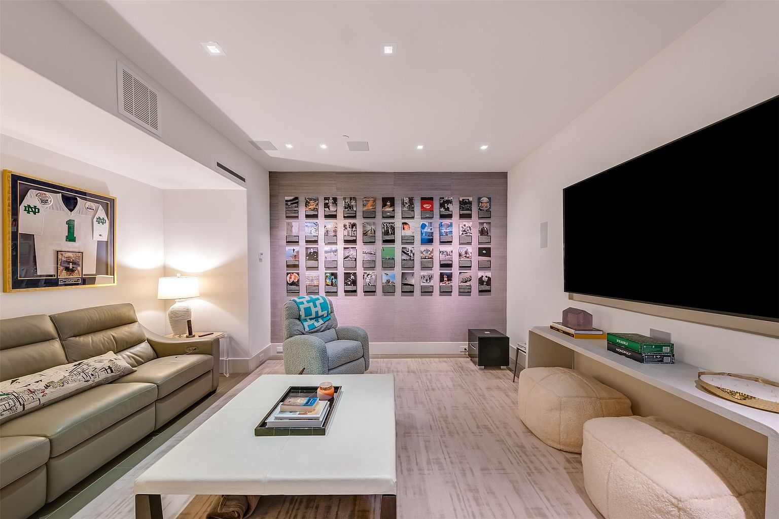 This contemporary media room features a comfortable leather sofa, a plush armchair, and two soft ottomans arranged around a central coffee table. The focal point is a large wall-mounted television positioned above a sleek console, while the opposite wall displays a curated gallery of framed prints. The space is illuminated by recessed ceiling lighting and a warm table lamp, creating a cozy and inviting atmosphere for entertainment.