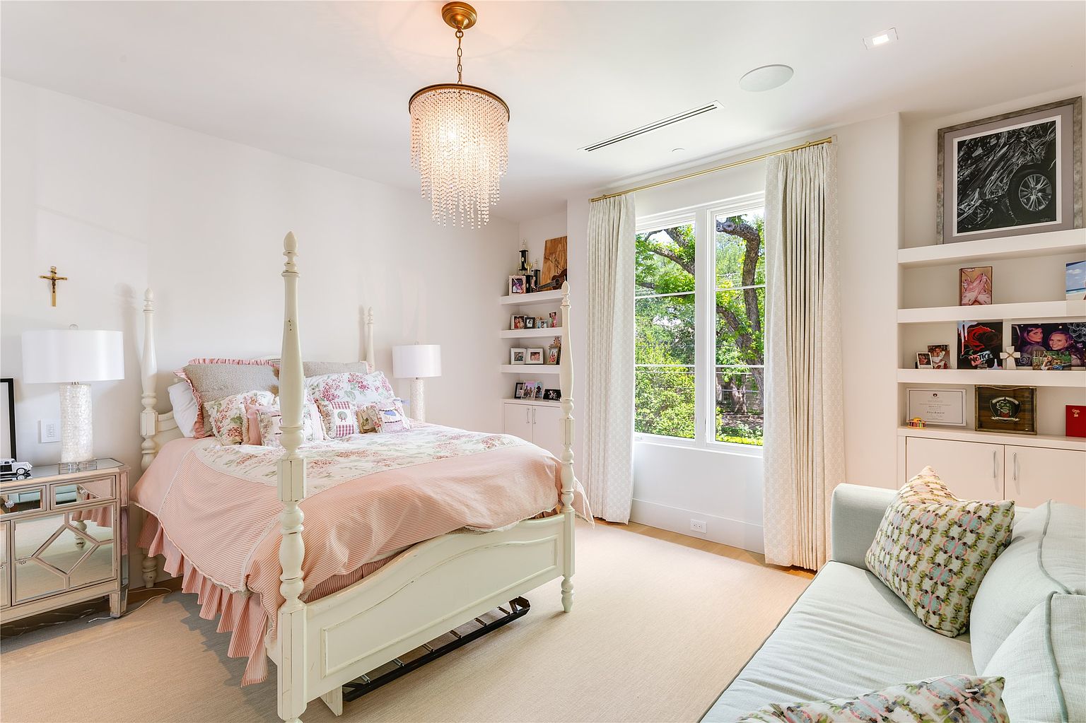 This bright and airy bedroom features a classic four-poster bed dressed in floral and striped linens, centered beneath a sparkling crystal chandelier. The room includes built-in shelving units flanking a large window that provides ample natural light and a view of the lush greenery outside. A light-colored sofa sits in the foreground, creating a cozy seating area that complements the soft, neutral color palette of the space.