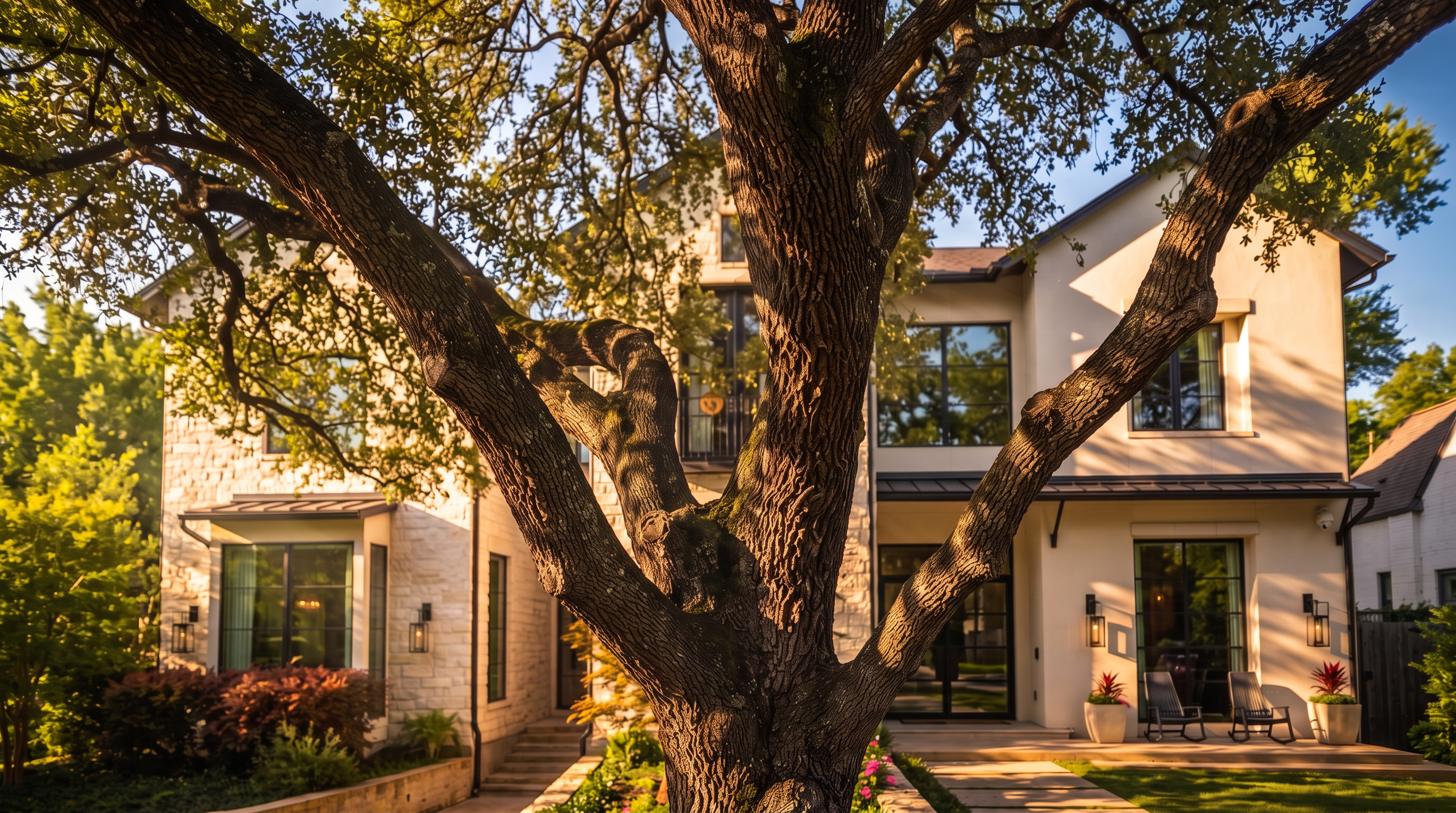 This stunning rear elevation of a luxury home features a combination of stone and smooth stucco finishes, highlighted by expansive black-framed windows that invite natural light. A prominent, mature tree in the foreground frames the house, while a beautifully landscaped terrace with comfortable outdoor seating creates an inviting space for relaxation. The design balances architectural sophistication with a serene, private backyard setting.