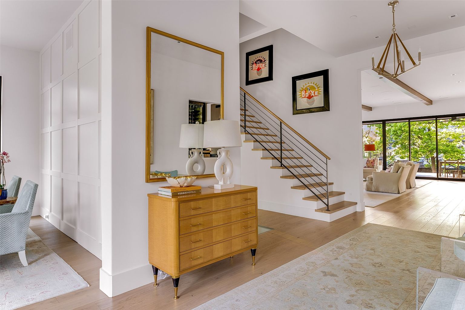 This bright and airy entryway features a modern staircase with a sleek metal railing and light wood treads. A warm-toned wooden chest sits beneath a large, gold-framed mirror, creating a welcoming focal point, while the open floor plan leads the eye toward a spacious living area with large glass doors. The space is characterized by clean white walls, light wood flooring, and a sophisticated, contemporary aesthetic.
