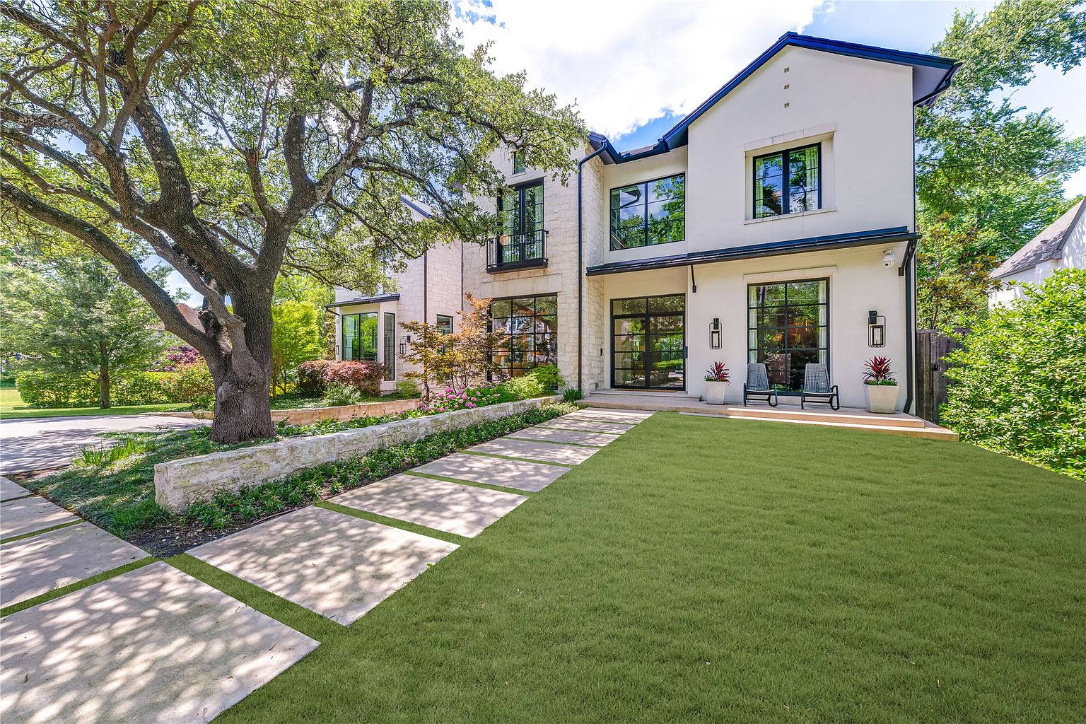 This striking modern residence features a sophisticated blend of white stucco and light-colored stone masonry, accented by dark-framed windows and doors. A wide, geometric paver walkway leads through a manicured lawn toward the entrance, which is framed by a large, mature oak tree and a low stone retaining wall. The composition offers a cinematic perspective of the home's clean lines and elegant landscaping, conveying a sense of luxury and curb appeal.