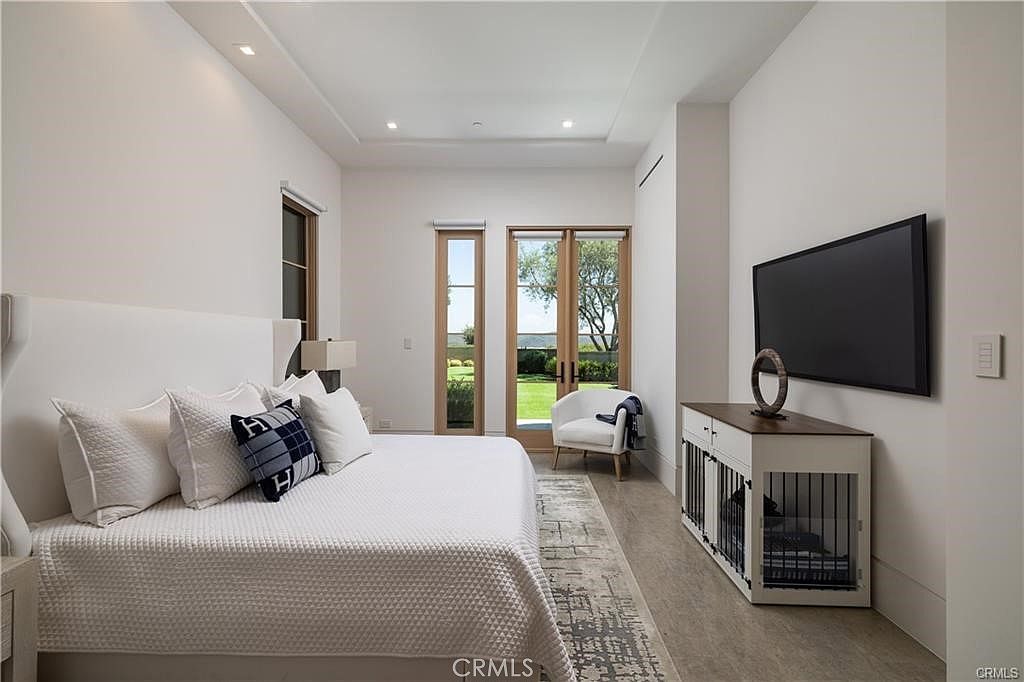 This spacious primary bedroom features a neutral color palette, light wood-toned flooring, and a large bed with a tall, upholstered headboard. A white media console with integrated pet crates sits beneath a wall-mounted television, while a pair of glass-paned doors provides a view of the outdoor greenery. The room is illuminated by recessed lighting and natural light, creating a serene and sophisticated atmosphere.