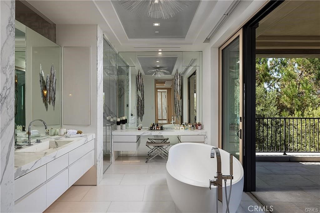 This luxurious primary bathroom features a sleek, white freestanding soaking tub positioned near large sliding glass doors that open to a private balcony. The space includes a long vanity with a stone countertop, a dedicated makeup vanity area with a stool, and expansive mirrors that enhance the sense of openness. The room is finished with light-toned tile flooring and modern, minimalist fixtures, creating a bright and sophisticated atmosphere.