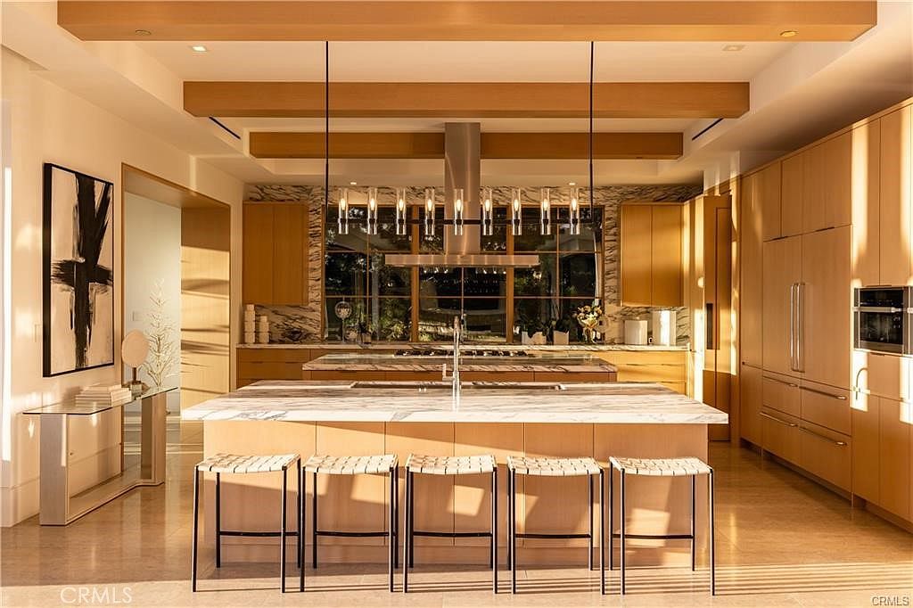 This spacious, modern kitchen features a large central island with a stone countertop and seating for five, complemented by light wood cabinetry and integrated appliances. A linear pendant light fixture hangs above the island, while exposed wooden beams add architectural interest to the ceiling. The space is bathed in warm, natural light, creating an inviting and sophisticated atmosphere.