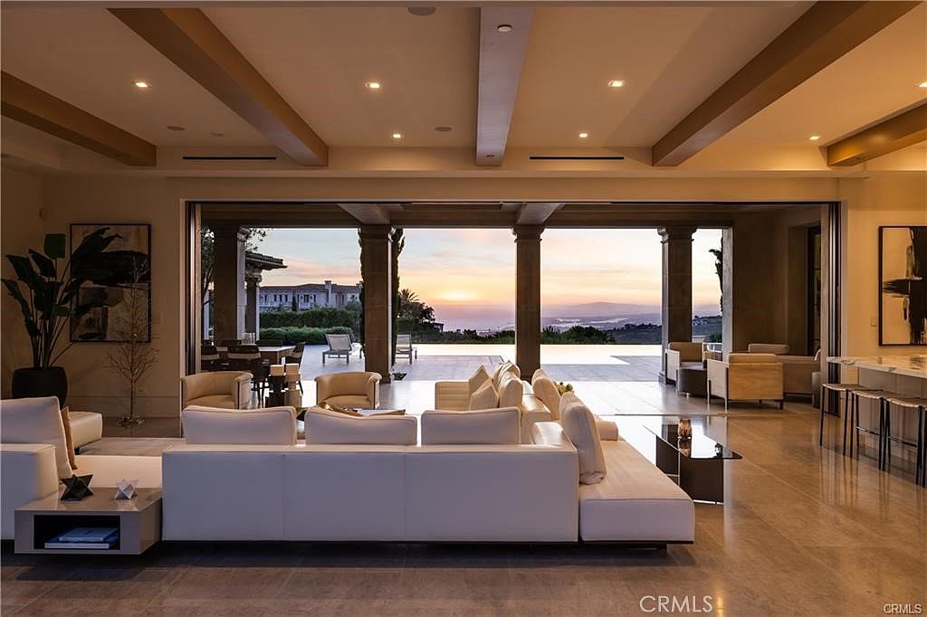 This expansive, open-concept living room features a large white sectional sofa and a neutral color palette that emphasizes a seamless transition to the outdoors. The space is defined by a series of large, retractable glass walls that open onto a covered patio, revealing a stunning sunset view over a pool and distant hills. The room is accented by warm wood-toned ceiling beams and recessed lighting, creating a sophisticated and airy atmosphere.