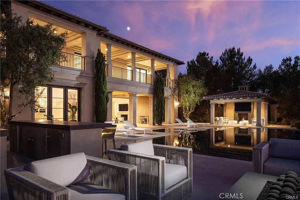 This luxurious backyard scene features a large swimming pool reflecting the twilight sky and the illuminated architecture of a multi-story Mediterranean-style home. The patio area is furnished with modern, light-toned outdoor seating and a built-in kitchen counter, while a separate covered pavilion with a fireplace sits across the water. The composition captures a serene, high-end evening atmosphere, emphasizing the seamless transition between indoor and outdoor living spaces.