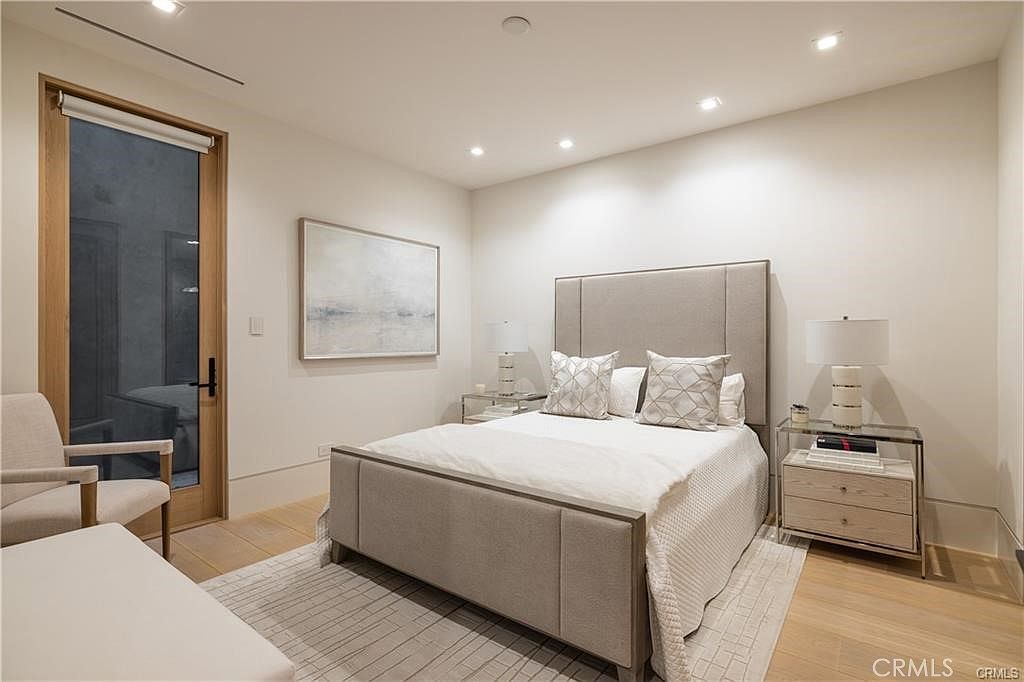 This contemporary bedroom features a neutral color palette with a large upholstered headboard as the focal point. The room is furnished with a bed flanked by two glass-topped nightstands with matching lamps, and a light-colored armchair sits in the corner. A tall glass door with a wood frame provides access to the outdoors, while recessed lighting and a textured area rug complete the clean, minimalist aesthetic.