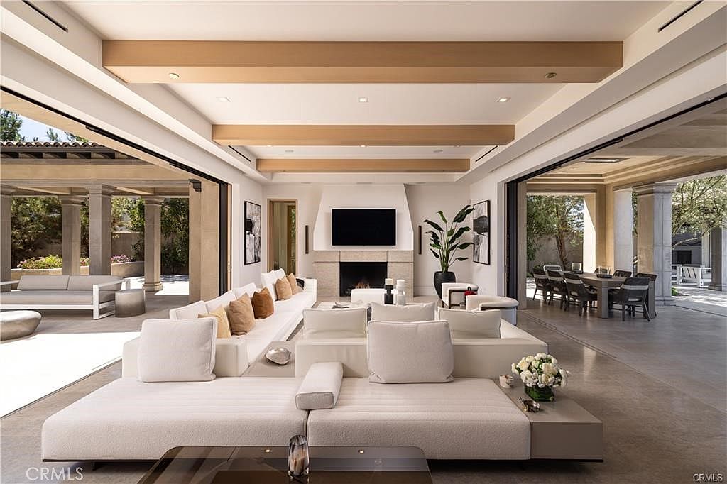 This expansive, open-concept living room features a neutral color palette and seamless indoor-outdoor flow provided by large sliding glass walls on both sides. The space is anchored by a central fireplace with a tall, light-toned surround and a mounted television, flanked by a potted plant. The room is furnished with a large, modular white sofa arrangement, light wood ceiling beams, and polished concrete flooring, creating a sophisticated and airy atmosphere.