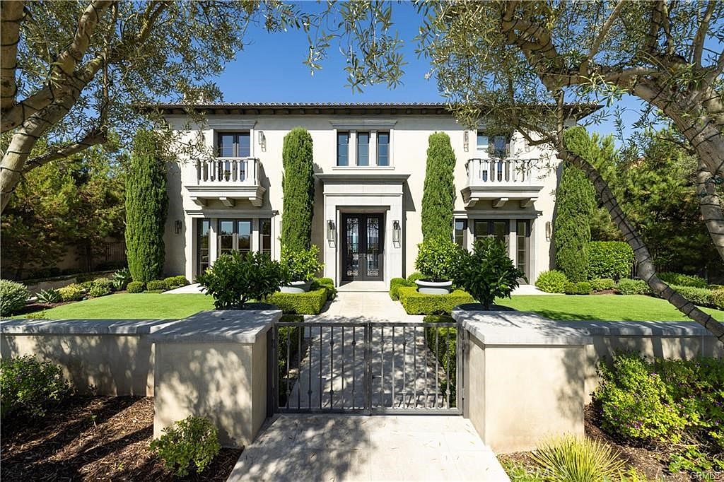 This elegant Mediterranean-style residence features a symmetrical facade with a central entryway, flanked by tall, manicured cypress trees and lush green lawns. The exterior is finished in a light-toned stucco, complemented by dark-framed windows and two symmetrical balconies on the second level. A stone-paved walkway leads through a decorative metal gate to the front door, creating a grand and inviting curb appeal.