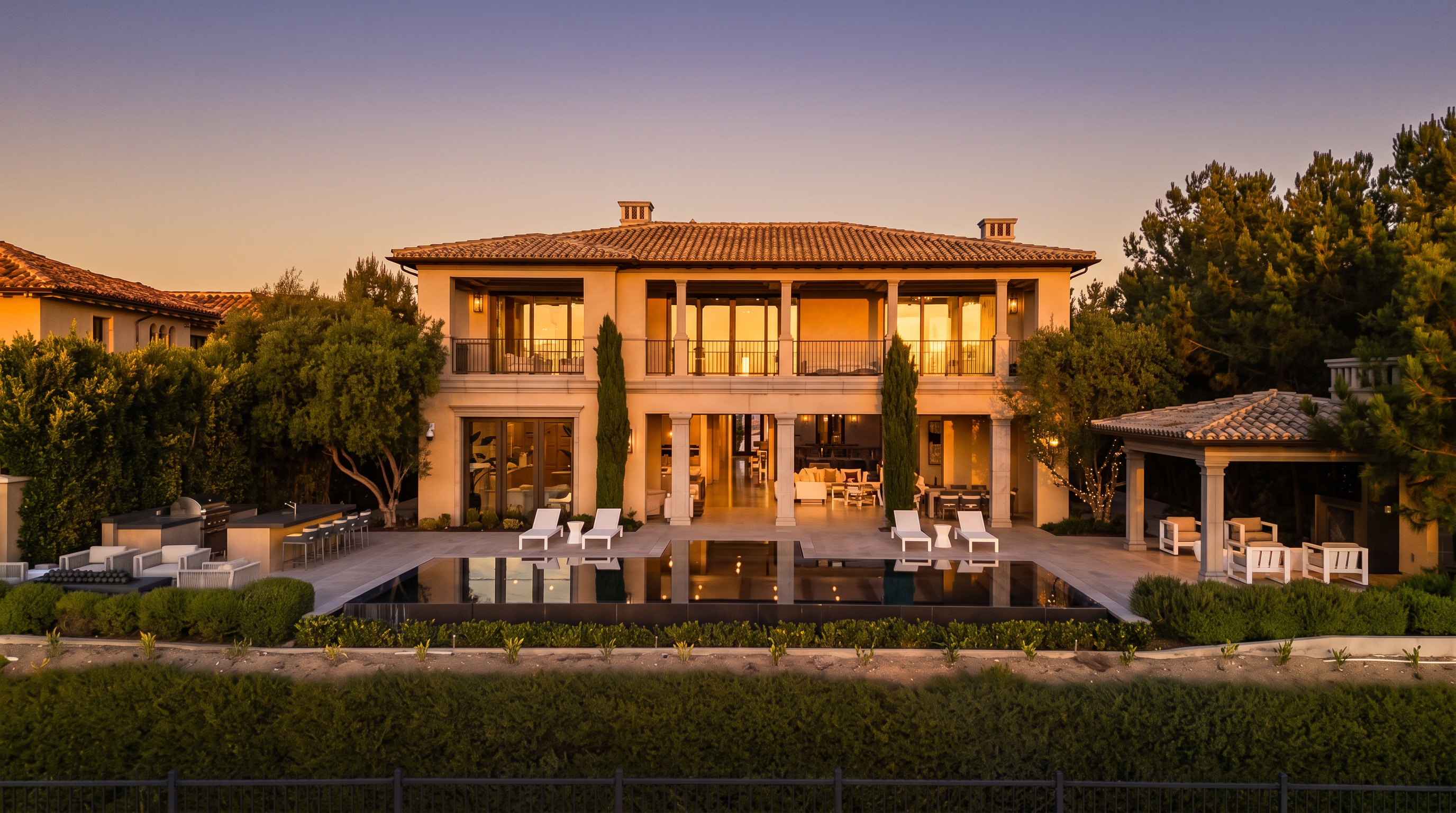 This elegant two-story Mediterranean-style estate is captured from a rear perspective, showcasing a symmetrical facade with a central loggia and a large, dark-tiled infinity pool. The outdoor living space includes a covered patio area with lounge seating, an outdoor kitchen with bar stools, and a separate poolside cabana. The property is framed by lush landscaping and mature trees, creating a serene and luxurious atmosphere at dusk.