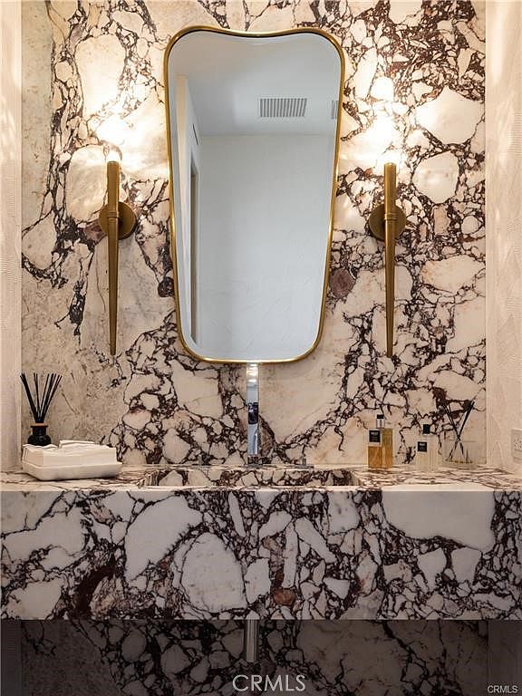 This elegant powder room features a striking vanity and wall treatment clad in a bold, dark-veined stone. A uniquely shaped, gold-framed mirror is centered above a sleek, wall-mounted faucet, flanked by two slender, modern gold sconces. The space exudes a sophisticated and high-end aesthetic, emphasizing luxury materials and refined lighting.