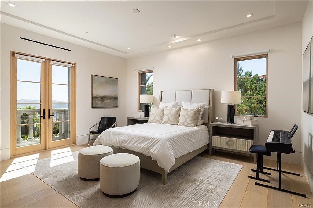 This bright and airy primary bedroom features a neutral color palette, light wood flooring, and a large bed with a tufted headboard. The room is accented by two round ottomans at the foot of the bed, a sleek black chair, and a digital piano, all set against a backdrop of soft white walls. Large glass doors open onto a balcony, allowing natural light to flood the space and providing a scenic view of the outdoors.