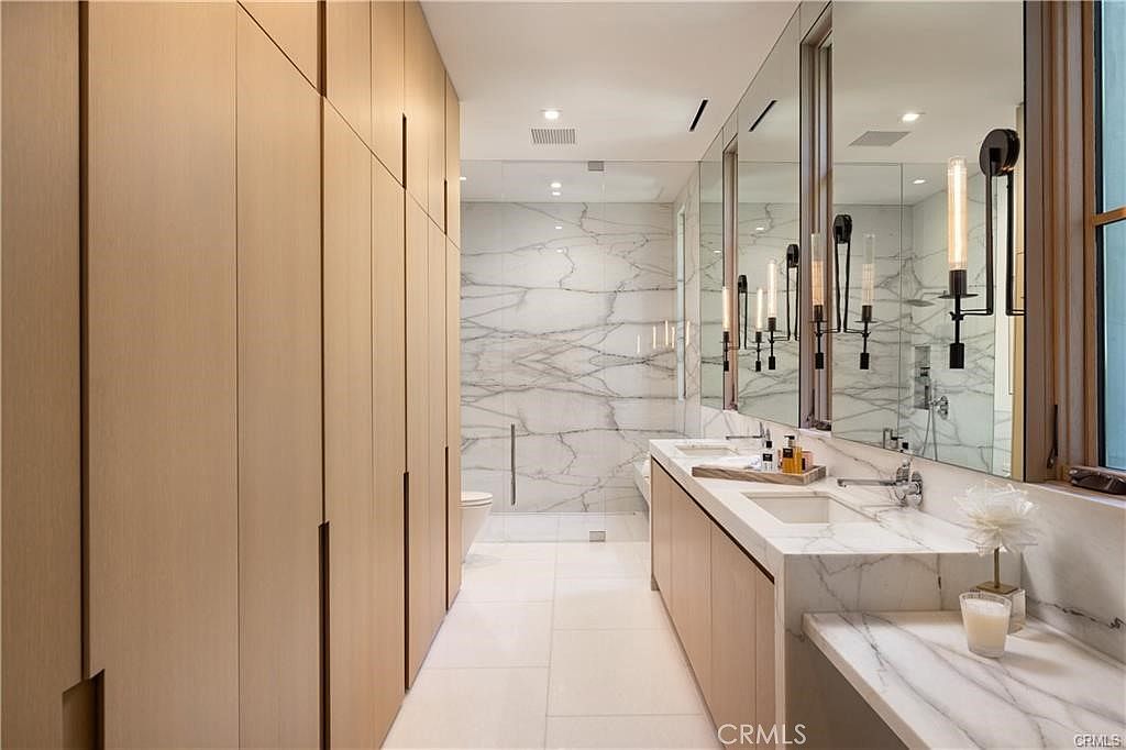 This modern primary bathroom features a sleek vanity with a stone countertop and dual sinks, complemented by tall, minimalist light fixtures mounted on large mirrors. A wall of light wood cabinetry provides extensive storage, while the back of the room showcases a walk-in shower enclosed by glass panels with a striking stone-patterned wall. The space is finished with light-colored tile flooring, creating a bright, clean, and high-end aesthetic.