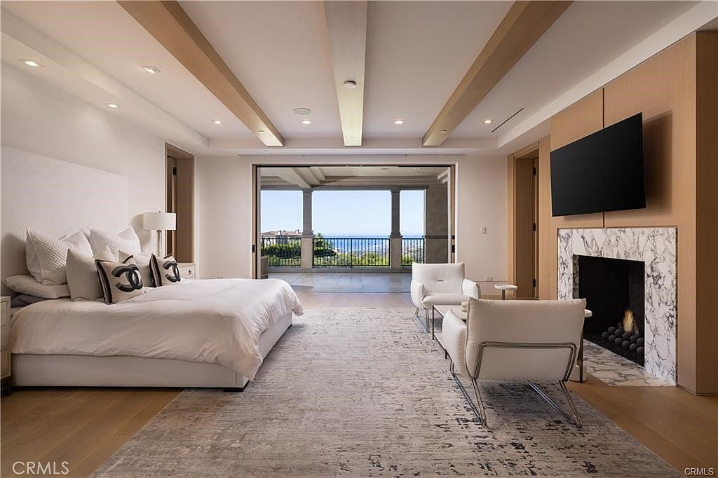This spacious primary bedroom features a neutral color palette, light wood flooring, and a large area rug. The room is anchored by a bed with white linens and a stone-clad fireplace surround with a mounted television, while large glass doors open to a balcony with ocean views. The ceiling is accented with exposed wooden beams, creating a sophisticated and airy atmosphere.
