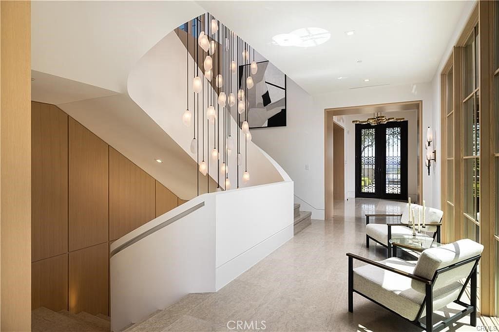 This elegant entryway and staircase area features a dramatic multi-pendant light fixture suspended from the ceiling, creating a striking focal point. The space is characterized by a clean, modern aesthetic with light-toned stone flooring, wood-paneled walls, and a minimalist white staircase railing. Two comfortable armchairs are positioned near the entrance, which leads to a dark-framed glass door, offering a bright and sophisticated first impression of the home.