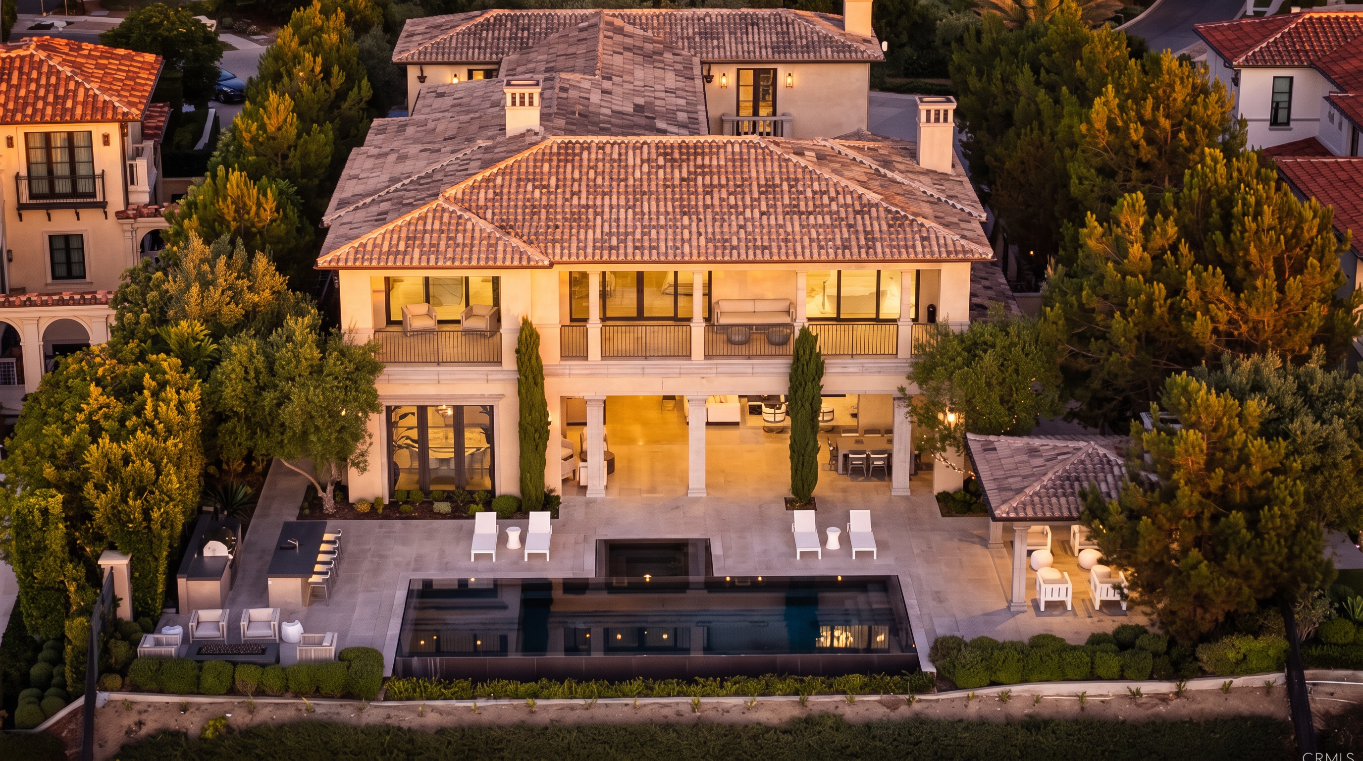 This aerial perspective captures the rear elevation of a luxurious multi-story residence at dusk, showcasing a symmetrical architectural design with a tiled roof and a spacious second-story balcony. The ground level features an expansive covered patio that transitions seamlessly to a sleek, rectangular swimming pool surrounded by stone decking and manicured landscaping. The scene is illuminated by warm interior and exterior lighting, creating an inviting and sophisticated atmosphere.