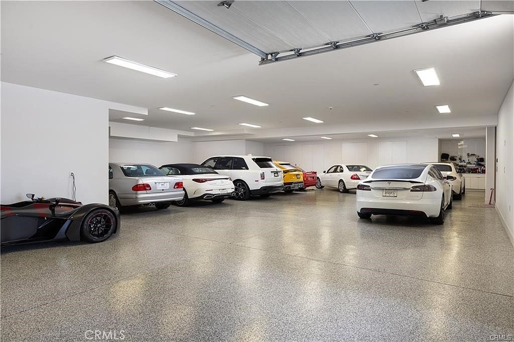 This expansive, well-lit garage features a polished, speckled epoxy floor and accommodates multiple luxury vehicles parked in a row. The space is finished with clean white walls and recessed ceiling lighting, creating a bright and professional atmosphere. The perspective captures the depth of the garage, showcasing the impressive scale and organized layout of this high-end storage area.