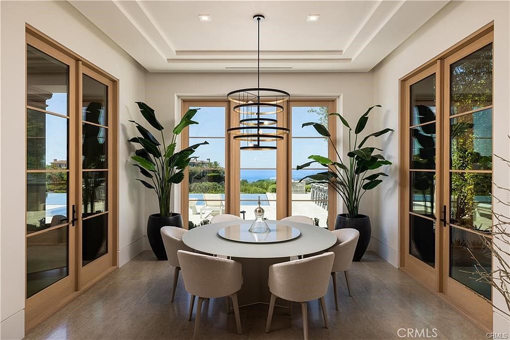 This elegant dining room features a central round table surrounded by six upholstered chairs, positioned beneath a modern, multi-tiered circular chandelier. The space is framed by large glass doors on either side and a central wall of windows that provide a clear view of the outdoor pool and coastal landscape. The room is accented by two large potted plants, creating a balanced and sophisticated atmosphere with a neutral color palette.
