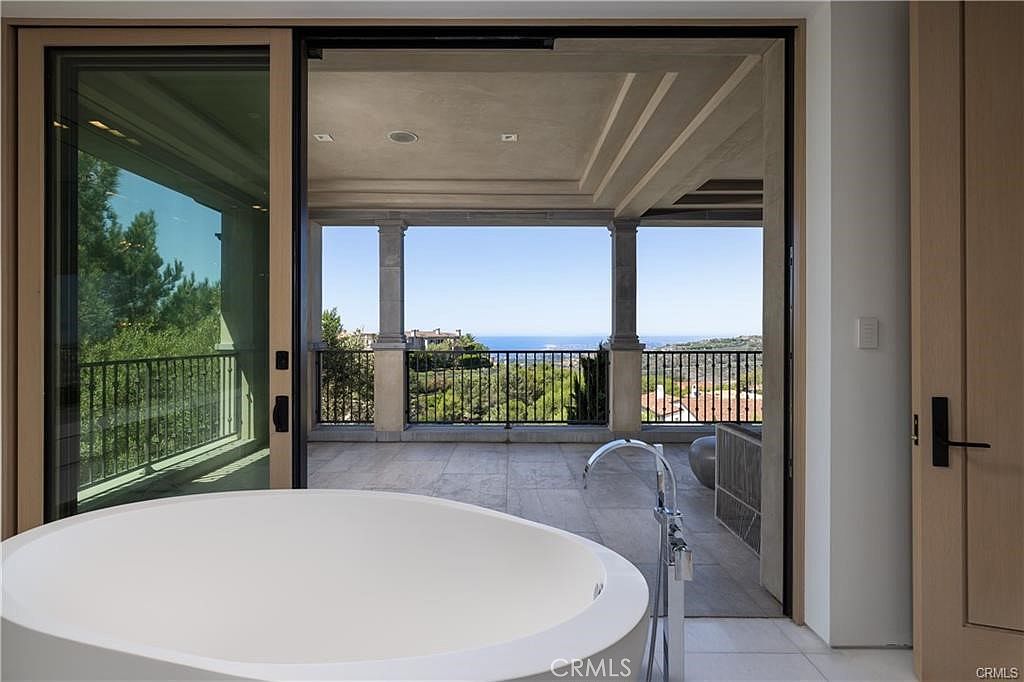 This primary bathroom features a large, white soaking tub positioned in front of expansive sliding glass doors that open onto a covered balcony. The balcony offers a scenic view of the surrounding hills and the distant ocean, framed by stone columns and a wrought-iron railing. The interior space is bright and minimalist, emphasizing a seamless transition between the indoor bathing area and the outdoor living space.