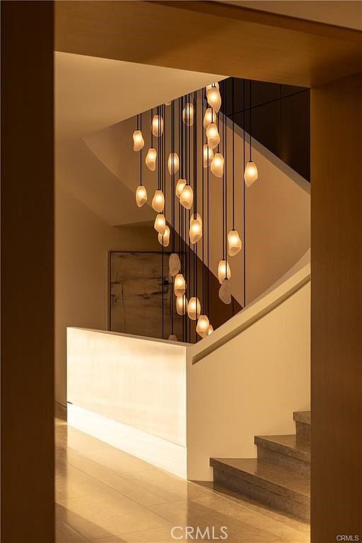 This interior view captures a dramatic staircase area illuminated by a striking, multi-pendant chandelier with organic, light-diffusing shades. The space features a clean, modern aesthetic with a minimalist white stair railing and stone steps, framed by warm-toned architectural elements. The perspective looks through a doorway, emphasizing the depth and artistic lighting design of the home's circulation space.