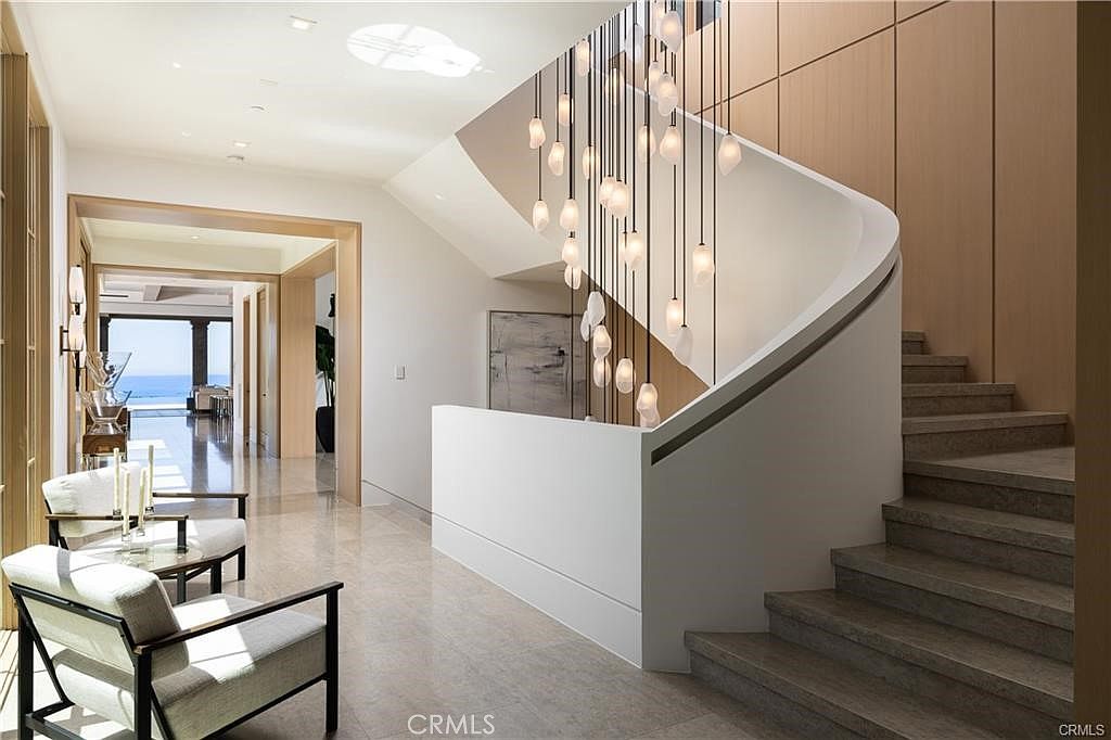 This elegant entryway features a dramatic, curved staircase with stone steps and a smooth, white balustrade. A striking, multi-pendant light fixture hangs from the ceiling, creating a focal point above the landing, while light-toned wood paneling adds warmth to the wall. The perspective looks through the foyer toward a bright, open hallway that leads to a view of the ocean in the distance.