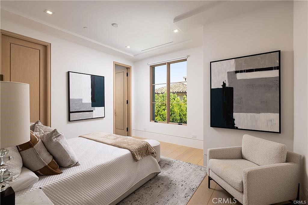 This bright and modern bedroom features light wood flooring, neutral walls, and a minimalist aesthetic. The room is furnished with a bed dressed in white linens, a textured throw, and a comfortable armchair, complemented by two large abstract art pieces on the walls. A window provides natural light and a view of the greenery outside, creating a serene and inviting atmosphere.