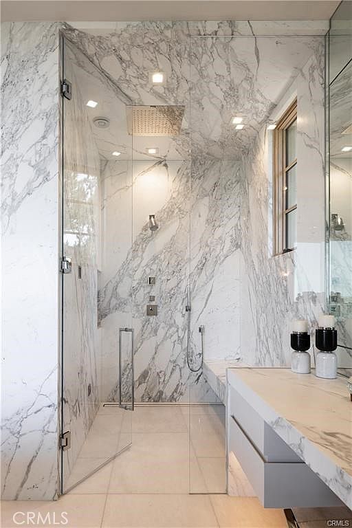 This luxurious bathroom features a walk-in shower enclosed by glass panels, showcasing extensive use of light-colored, heavily veined stone on the walls and vanity. The shower includes both a rainfall showerhead and a handheld sprayer, while a floating vanity with drawers provides a sleek, modern aesthetic. The perspective is from the doorway, capturing the seamless integration of the shower area with the vanity space.