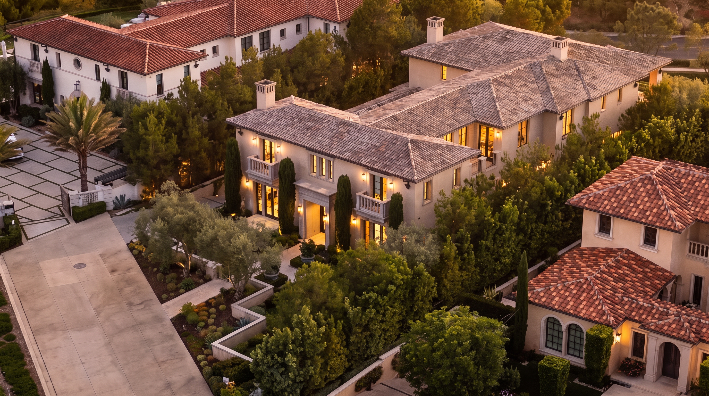 This high-angle aerial view captures a sprawling Mediterranean-style estate at dusk, showcasing its complex multi-gabled roofline with terracotta tiles and light-colored stucco walls. The property features a manicured landscape with mature trees, structured garden beds, and a paved driveway leading to the main entrance. The warm glow from the interior lighting highlights the architectural details, including balconies and arched windows, creating an inviting and luxurious atmosphere.