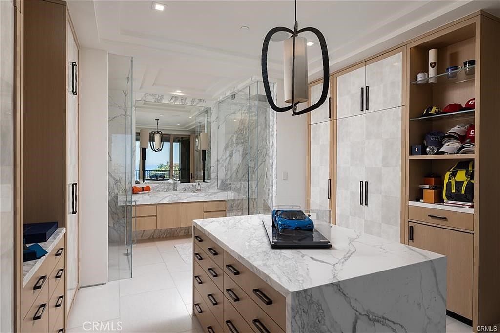 This luxurious walk-in closet features a central island with a stone countertop, showcasing a model car display. The space is outfitted with light wood cabinetry, open shelving for accessories, and a large mirror that reflects the adjacent bathroom area. A modern, sculptural pendant light hangs above the island, creating a sophisticated and organized atmosphere.