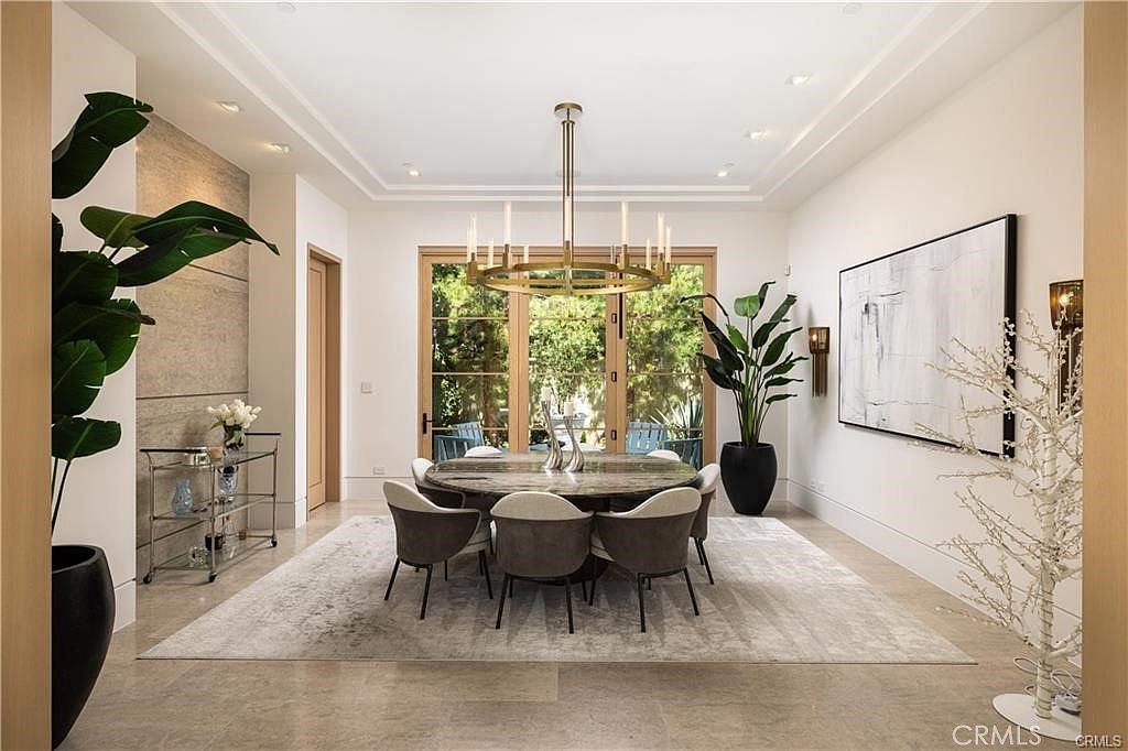 This elegant dining room features a large circular table surrounded by six modern, upholstered chairs, all centered on a textured area rug. A striking gold-toned chandelier hangs above the table, while a large abstract painting adorns the wall to the right. The space is framed by tall glass doors that provide a view of the lush greenery outside, creating a bright and sophisticated atmosphere.
