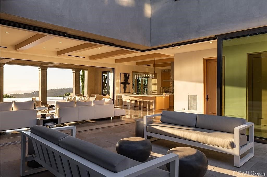 This expansive, open-concept living area seamlessly blends indoor and outdoor spaces through large sliding glass walls that open to a scenic terrace. The room features a contemporary design with light wood ceiling beams, neutral-toned sofas, and a view into a bright, modern kitchen with a central island. The warm, golden-hour lighting creates an inviting and luxurious atmosphere, highlighting the clean lines and sophisticated architectural flow of the home.