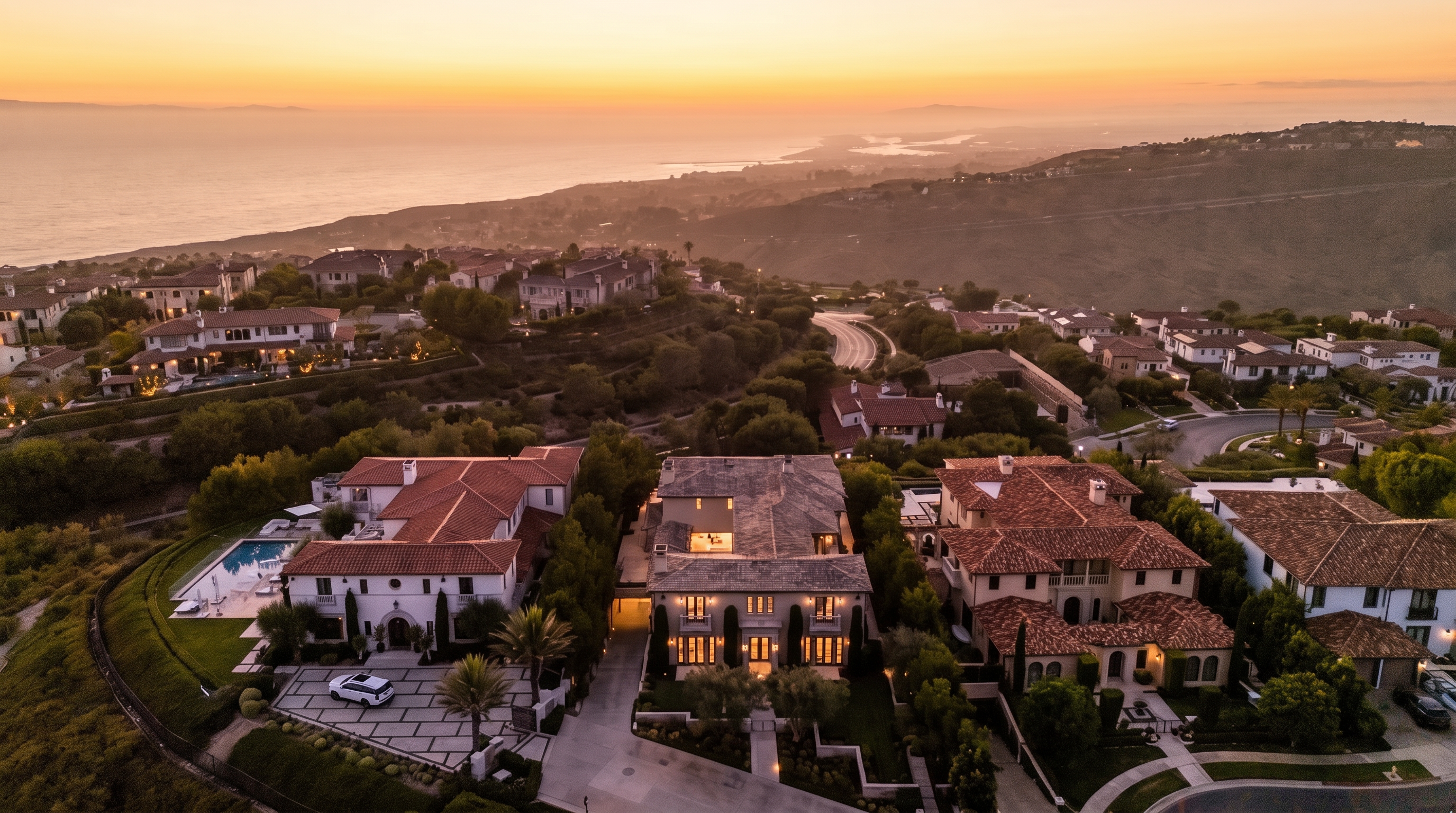 This high-angle aerial view captures a cluster of luxury Mediterranean-style homes nestled on a hillside overlooking a vast coastline at sunset. The scene highlights the architectural details of the properties, including terracotta-tiled roofs, paved driveways, and manicured landscaping, all set against a panoramic backdrop of the ocean and distant hills. The warm, golden light of dusk enhances the serene and upscale atmosphere of this coastal residential community.