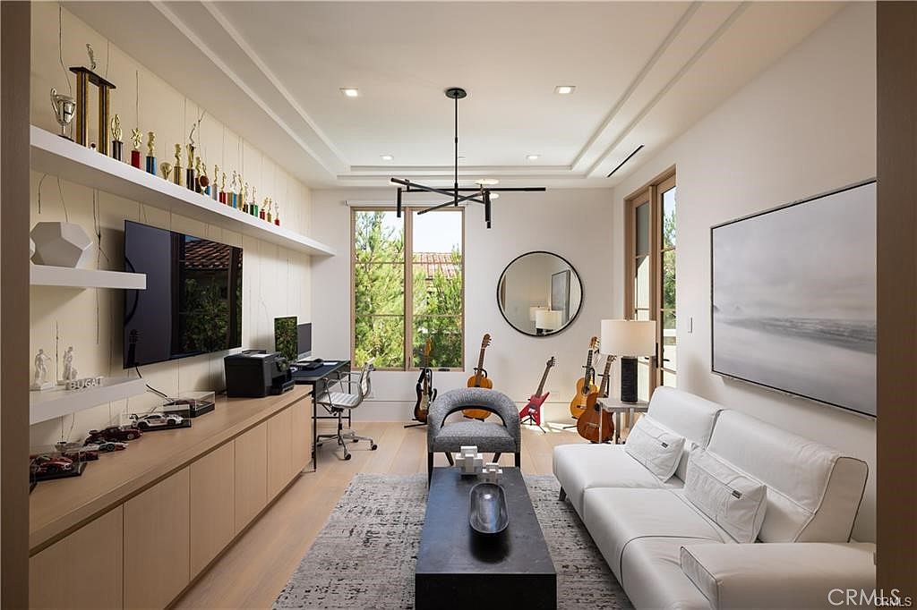 This sophisticated home office and media room features a sleek, light wood built-in desk and cabinetry unit with open shelving displaying trophies and collectibles. A white leather sofa faces a dark rectangular coffee table, while the room is accented by a collection of guitars and a large circular wall mirror. The space is illuminated by a modern geometric chandelier and large windows that provide a view of the surrounding greenery.