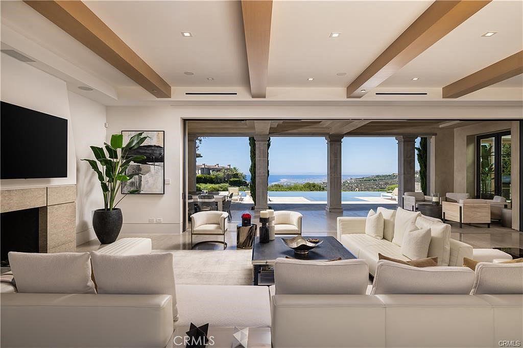This expansive, open-concept living room features a neutral color palette and high ceilings accented with light wood beams. The space seamlessly transitions to an outdoor patio through large, retractable glass doors that reveal a stunning ocean view and a swimming pool. The room is furnished with plush white sofas, a dark coffee table, and a tall stone-clad chimney breast, creating a sophisticated and airy atmosphere.