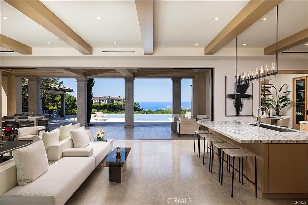 This expansive open-concept living and kitchen area features a seamless transition to an outdoor patio through large, retractable glass walls. The kitchen is anchored by a substantial island with a stone countertop and bar seating, while the adjacent living space is furnished with a modern white sofa. The room is defined by exposed wood ceiling beams and a neutral color palette, creating a bright, airy atmosphere that highlights the panoramic ocean view.
