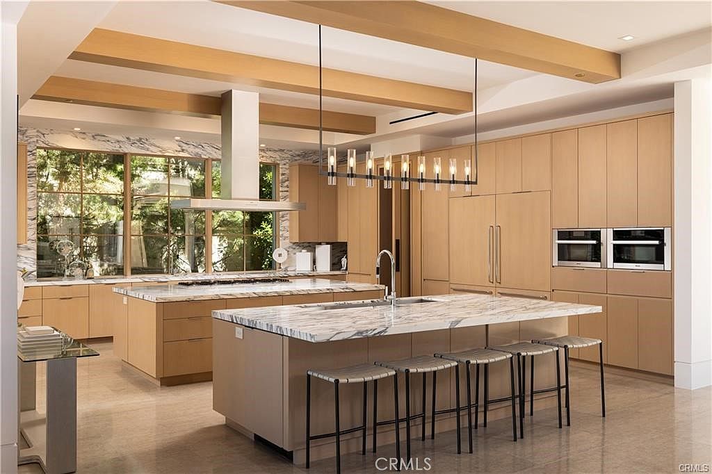 This expansive, modern kitchen features dual islands with light-toned stone countertops and cabinetry, creating a sophisticated and airy atmosphere. The space is defined by light wood cabinetry, a linear pendant light fixture, and large windows that provide a view of the lush greenery outside. The perspective is a wide-angle shot from the kitchen island, showcasing the seamless integration of high-end appliances and clean, minimalist design.