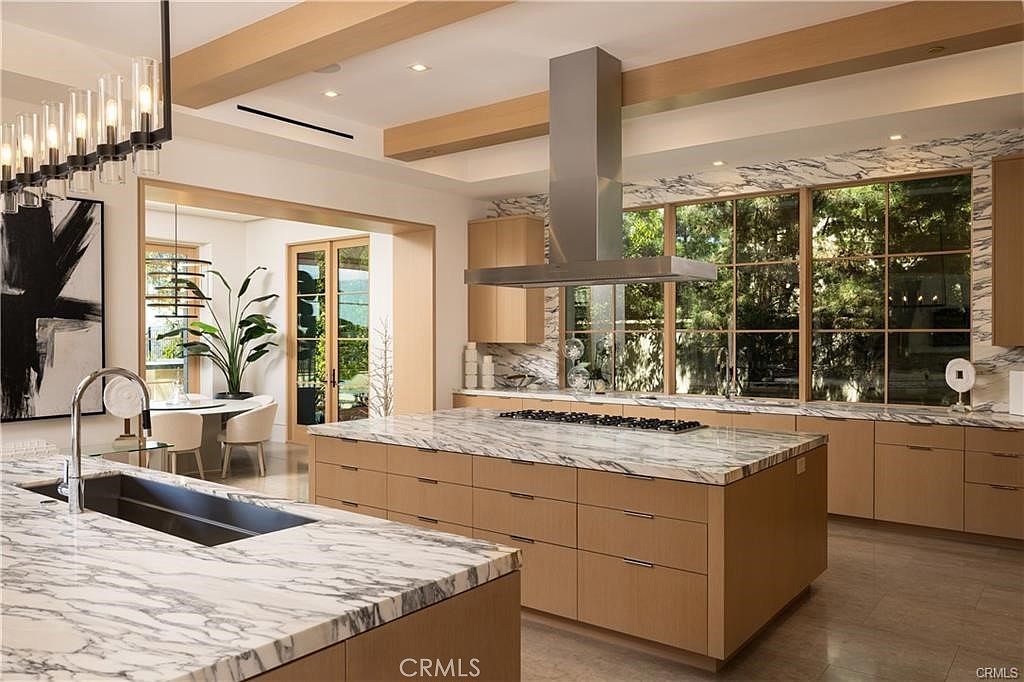 This expansive, modern kitchen features a large central island with a stone countertop and light wood cabinetry, complemented by a matching stone-topped peninsula in the foreground. A stainless steel range hood hangs above the cooktop, while large windows provide a bright, airy view of the lush greenery outside. The space is defined by clean lines, warm wood tones, and a sophisticated, open-concept design that flows into an adjacent dining area.