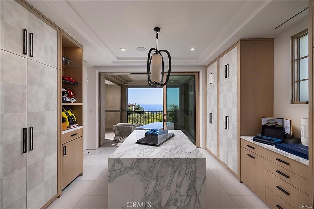 This sophisticated walk-in closet features a central stone-topped island with a display case, surrounded by custom light wood cabinetry with patterned paneling. The space is illuminated by a unique sculptural pendant light and offers a seamless transition to an outdoor balcony through large sliding glass doors. The overall aesthetic is modern and luxurious, emphasizing high-end finishes and an open, airy layout.