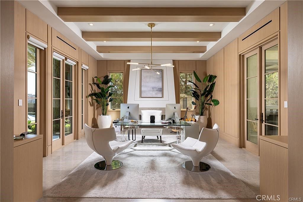 This sophisticated home office features light wood paneling on the walls and a coffered ceiling with exposed beams. A glass-topped desk sits at the center of the room, flanked by two modern white swivel chairs, while a tall, light-colored fireplace surround serves as a focal point between two large windows. The space is accented by two large potted plants and a neutral area rug, creating a professional yet serene atmosphere.
