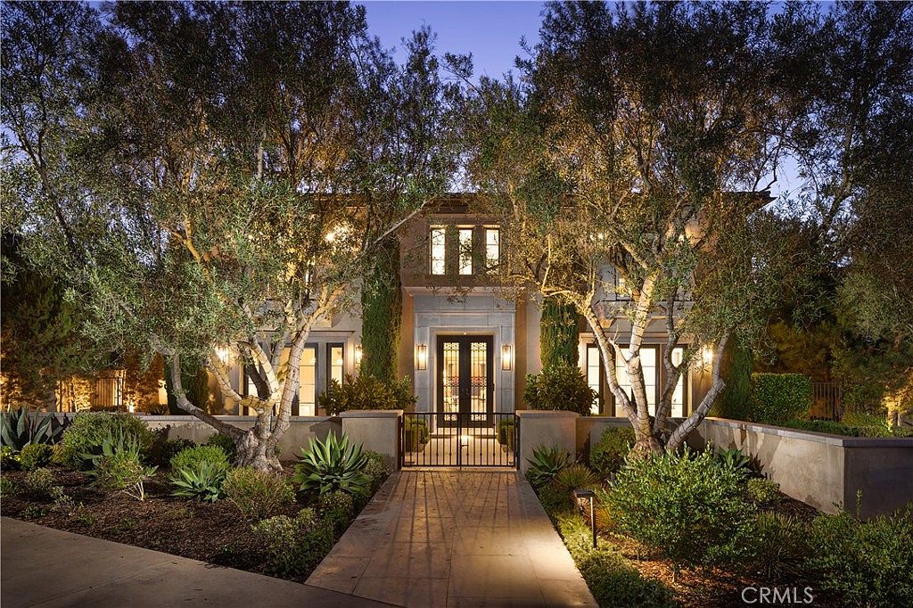 This elegant two-story residence is captured at dusk, showcasing a symmetrical facade framed by mature, illuminated olive trees. A stone-paved walkway leads through a decorative wrought-iron gate to the grand front entrance, which features a prominent stone surround and double doors. The warm exterior lighting highlights the architectural details and landscaping, creating an inviting and sophisticated curb appeal.