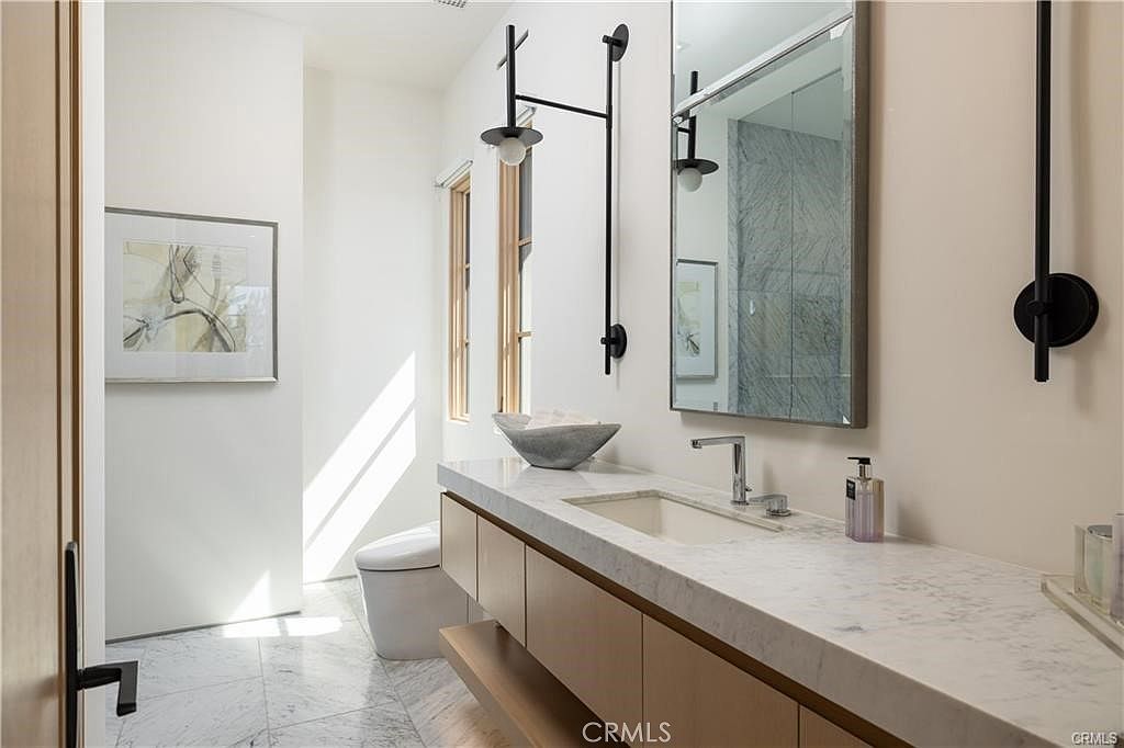 This modern bathroom features a floating vanity with light wood cabinetry topped by a stone counter and a vessel sink. A large rectangular mirror hangs above the vanity, flanked by sleek, black wall-mounted light fixtures. The space is finished with light-colored stone flooring and white walls, creating a bright and minimalist aesthetic.