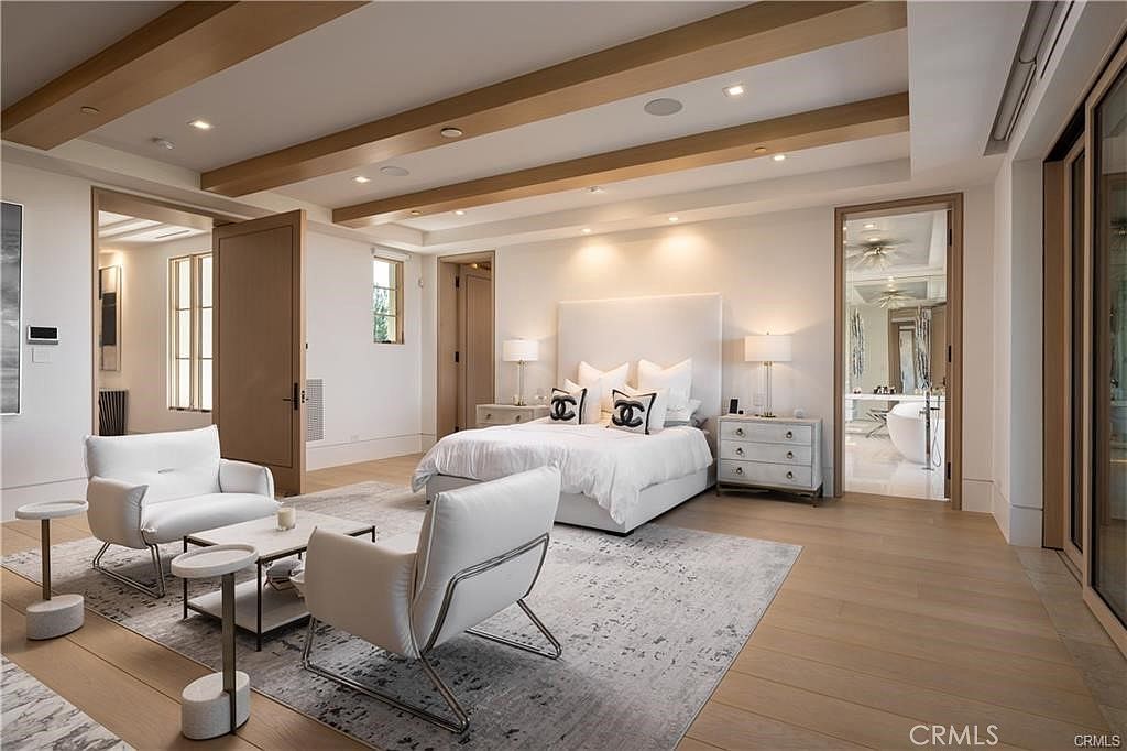 This spacious primary bedroom features a neutral color palette with light wood flooring and exposed ceiling beams. The room is furnished with a central bed flanked by matching nightstands, a seating area with two white armchairs, and a coffee table, all set upon a large patterned area rug. A doorway leads to an adjacent bathroom, while the overall design exudes a clean, modern, and luxurious aesthetic.