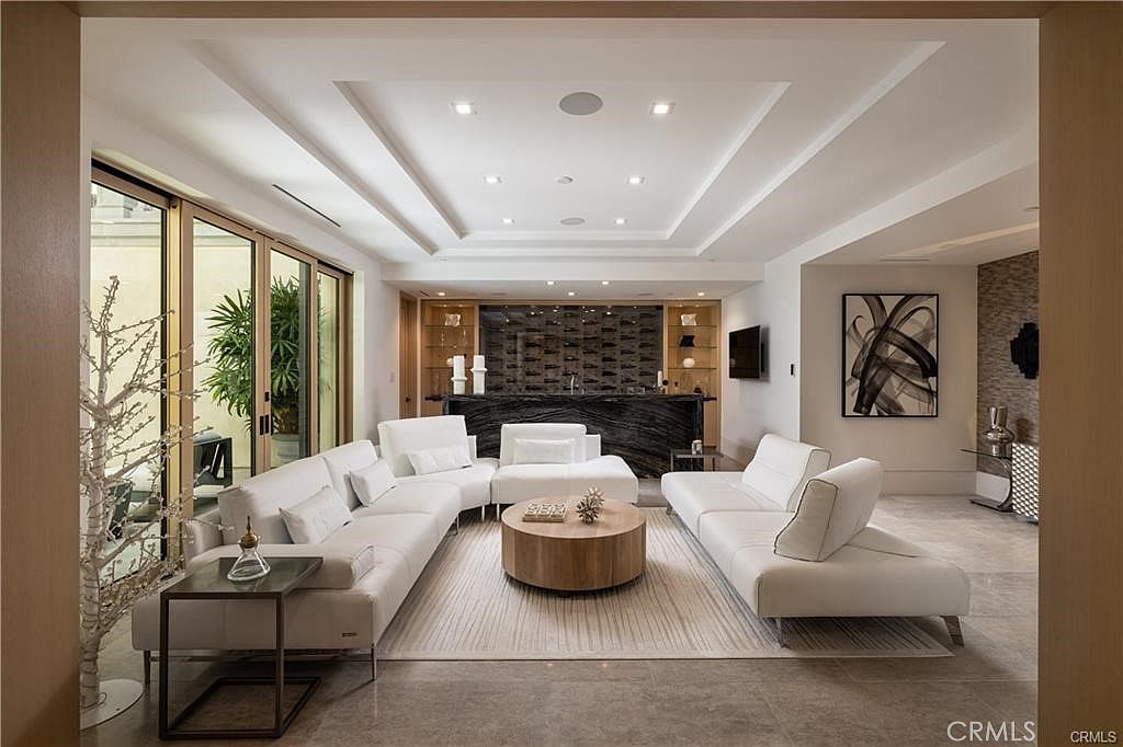This sophisticated living room features a spacious open-concept layout with a large white sectional sofa arranged around a circular light wood coffee table. In the background, a sleek bar area with a dark stone counter and integrated wine storage serves as a focal point, complemented by recessed ceiling lighting and a neutral color palette. Large glass sliding doors on the left provide a seamless transition to an outdoor space, while a piece of abstract wall art adds a modern touch to the refined interior.