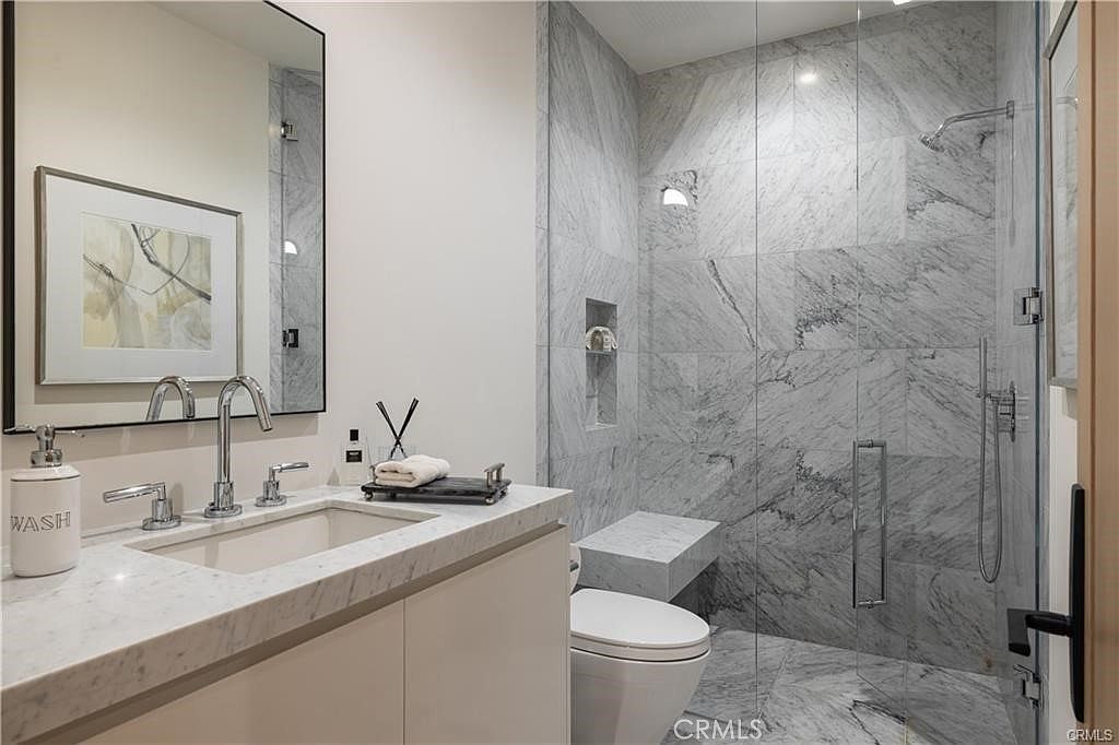This modern bathroom features a sleek vanity with a stone countertop and a rectangular undermount sink, complemented by a large mirror and minimalist fixtures. To the right, a glass-enclosed walk-in shower showcases light-toned stone wall tiles and a built-in stone bench. The space is bright and contemporary, with a toilet positioned neatly between the vanity and the shower area.