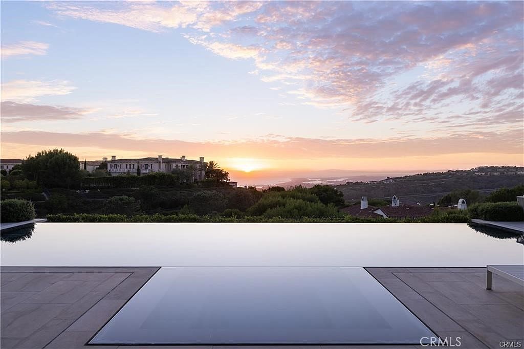 This image captures a stunning infinity-edge pool overlooking a vast, rolling landscape at sunset. The foreground features a sleek, tiled pool deck leading to the water's edge, which seamlessly blends into the horizon. In the distance, a large estate sits atop a hill, framed by a vibrant, cloud-filled sky that transitions from soft pinks to deep blues.