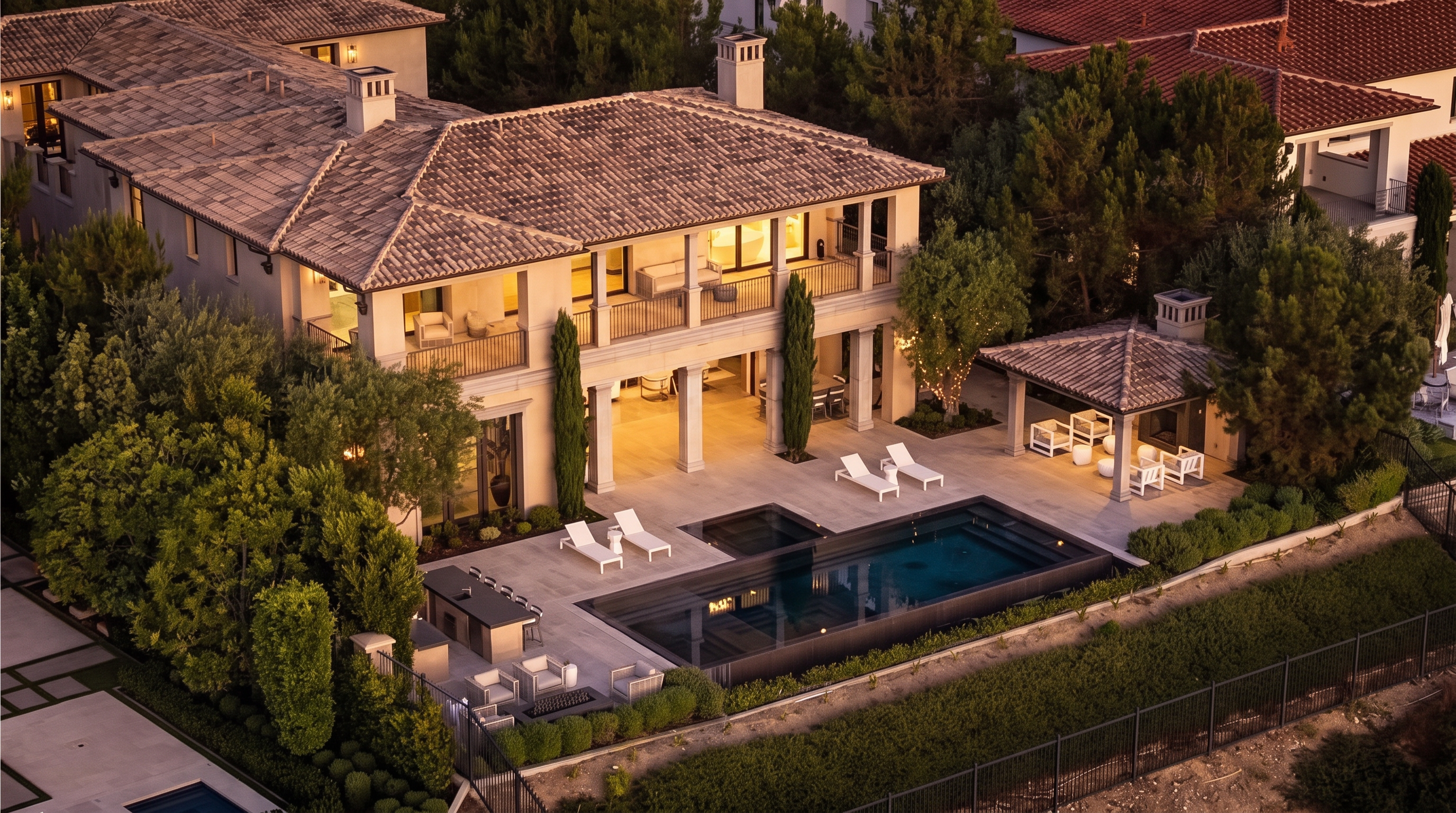 This high-angle aerial view captures a luxurious Mediterranean-style estate at dusk, highlighting its expansive backyard and pool area. The property features a multi-level structure with a tiled roof, a covered patio with outdoor seating, and a sleek rectangular swimming pool surrounded by stone decking. Lush landscaping, including tall cypress trees and manicured hedges, frames the outdoor living space, creating a private and sophisticated atmosphere.