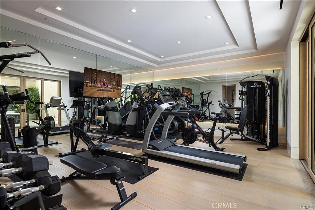 This professional-grade home gym features a wide array of fitness equipment, including a treadmill, rowing machine, stationary bike, and a multi-station strength training system. The room is enhanced by large wall-to-wall mirrors that create a sense of depth, while a flat-screen television is mounted for entertainment during workouts. Light wood flooring and recessed ceiling lighting provide a bright, clean, and motivating atmosphere for exercise.