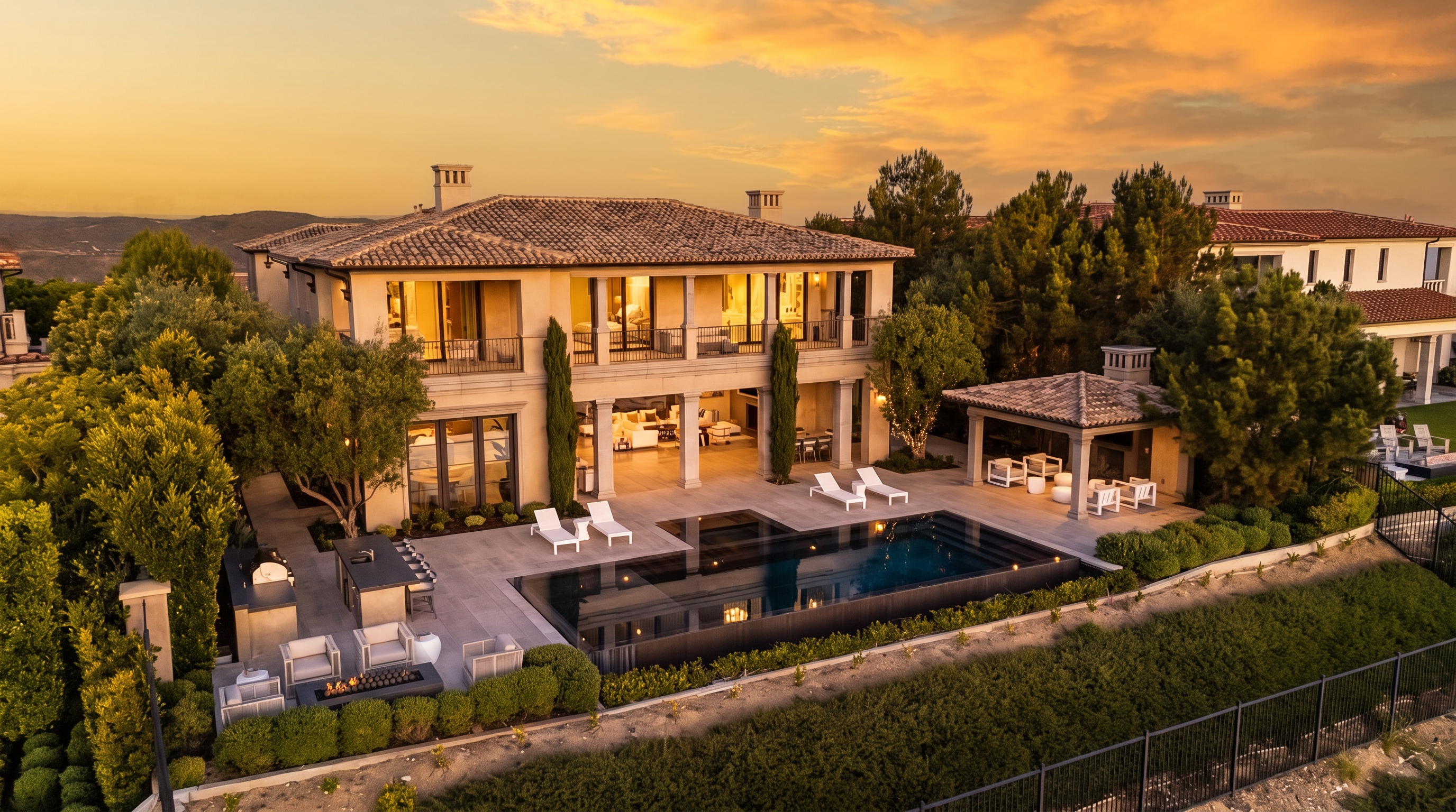 This aerial perspective captures a luxurious Mediterranean-style estate at dusk, highlighting a multi-level residence with a tiled roof and expansive balconies. The backyard features a sleek, dark-bottomed infinity pool, a covered outdoor lounge area with a stone-clad chimney breast, and a spacious stone patio perfect for entertaining. Lush landscaping and mature trees frame the property, creating a serene and private atmosphere.
