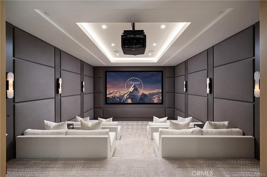 This image showcases a sophisticated home theater room designed for an immersive cinematic experience. The space features tiered seating with plush white sofas, dark upholstered wall panels, and a large projection screen centered on the back wall. The room is illuminated by recessed ceiling lights and wall-mounted sconces, creating a moody and luxurious atmosphere.